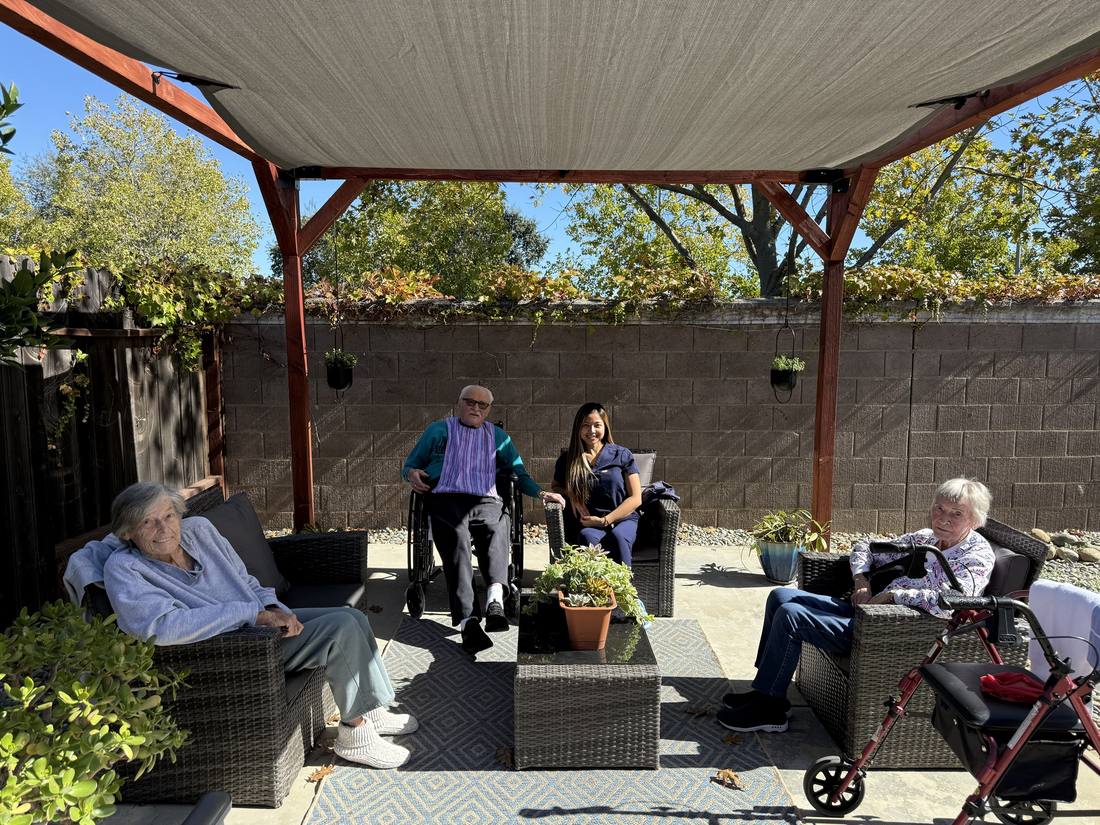 Three residents and a caregiver relaxing under a shaded patio pergola