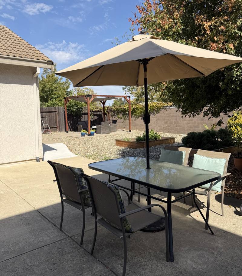 Backyard patio table under an umbrella, with the shaded pergola, garden beds and autumn leaves behind